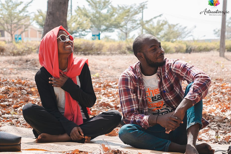 Image of a girl in a head scarf, laughing as she holds up her hand to her chest and a man, also smiling. both subjects are sitting on a mat